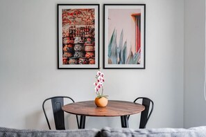 Ein Essbereich mit einem runden Tisch, zwei Stühlen und einer Vase mit Blumen. Darüber an der Wand hängen zwei gerahmte Kunstwerke.