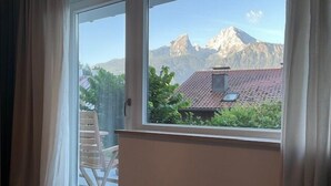 Una habitación con puerta de vidrio corrediza que da a un balcón. El balcón ofrece vistas a un paisaje montañoso con una casa de techo de tejas rojas.