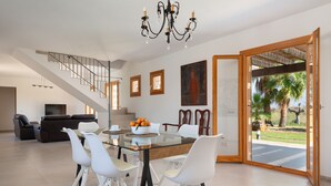 Un vaste espace de vie comprenant une table à manger en verre, des chaises blanches, un escalier et une grande fenêtre donnant sur des palmiers.