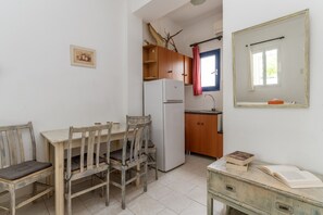 Een kleine keuken met een koelkast, houten kastjes, een eettafel met stoelen en een spiegel aan de muur.