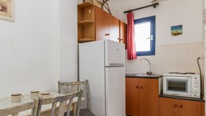 A compact kitchen with a refrigerator, microwave, wooden cabinets, a window with a curtain, and a small table with cups. - Holiday Home '9 Perasma Kypri Beach' with Private Terrace, Wi-Fi and Air Conditioning (Kypri)