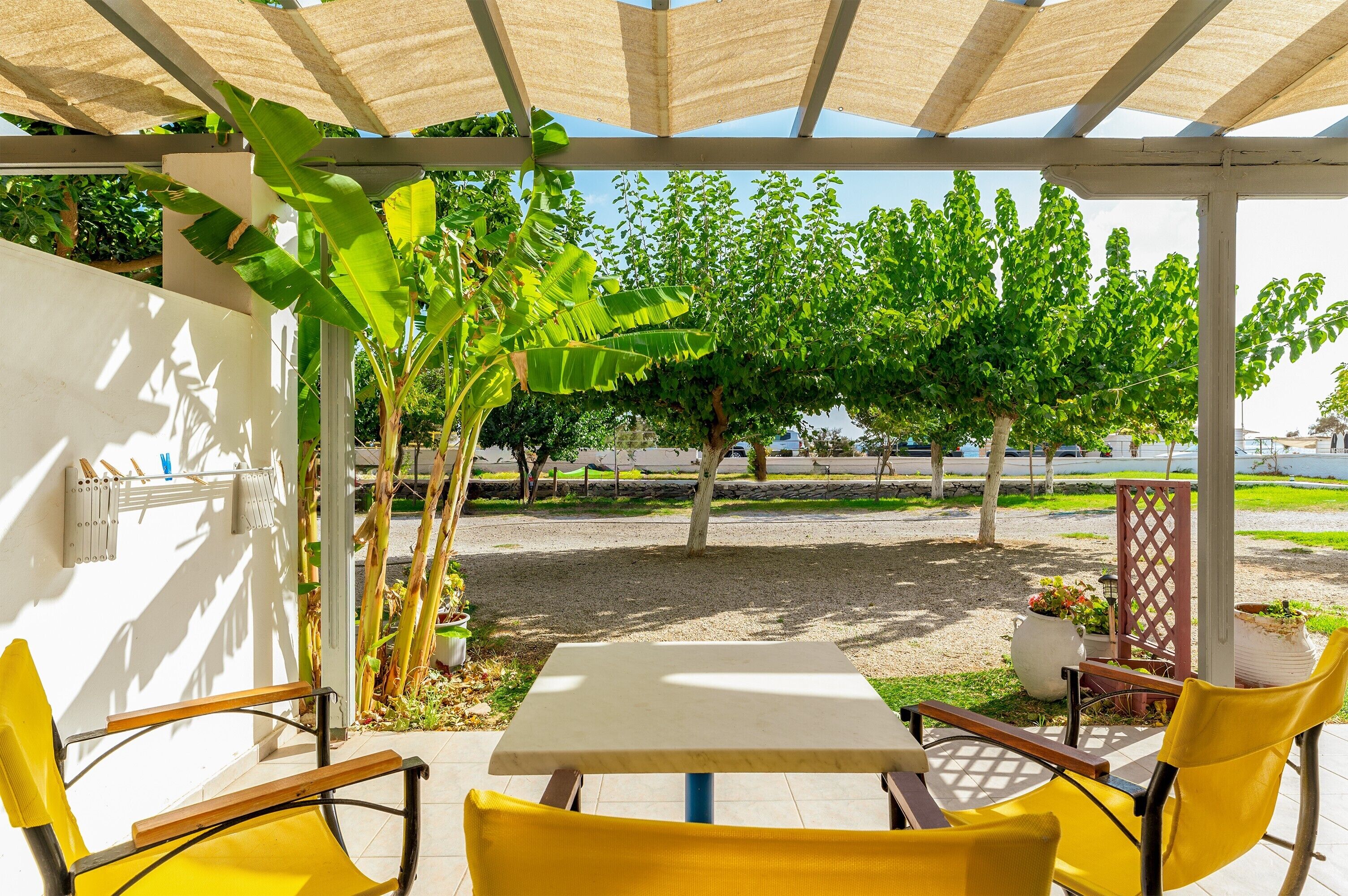 Een overdekt terras met een tafel en gele stoelen, omgeven door groen en bomen.