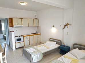 A compact hotel room with a kitchenette, two single beds, a small table, and a nightstand.