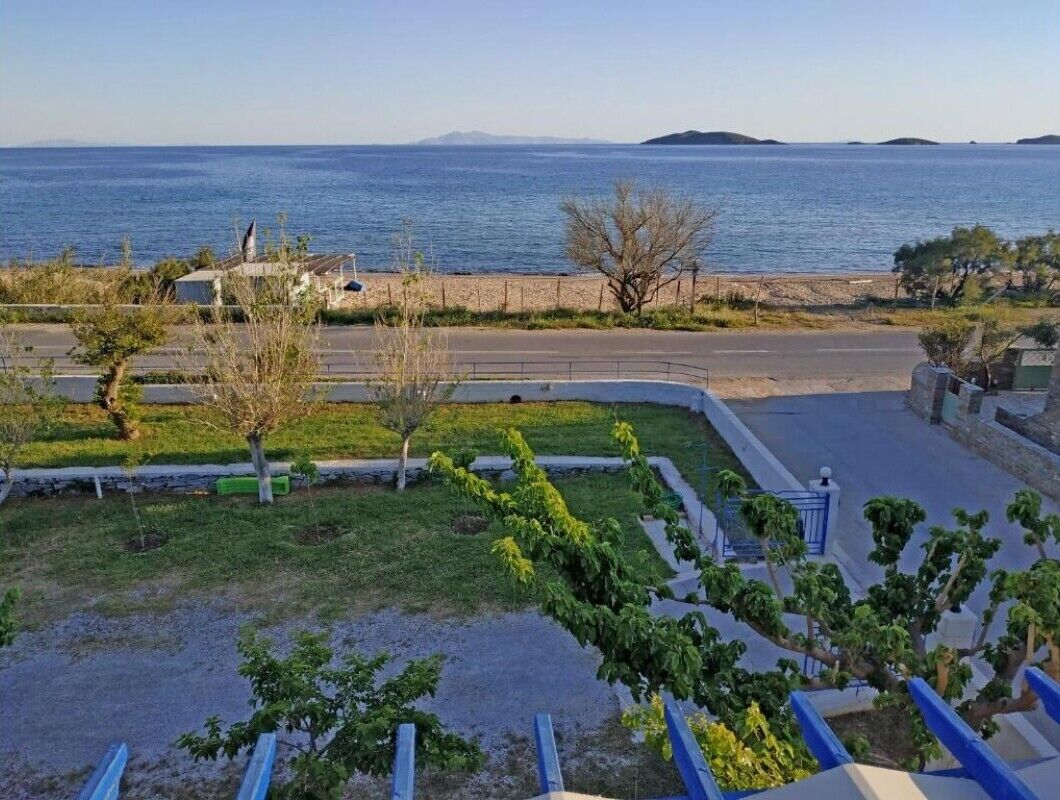A coastal residential area with a clear view of the sea, a paved road, and a grassy area with trees.
