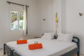 A single bed with white linens, two orange towels, a nightstand with a book, and a window with a curtain.
