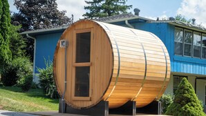 A wooden sauna structure with a door, placed on a wooden platform in front of a blue building.