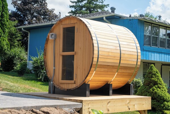 Una estructura de sauna de madera con una puerta, ubicada sobre una plataforma de madera frente a un edificio azul.