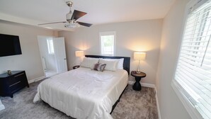 A bedroom with a large bed, two bedside lamps, a ceiling fan, a TV on the wall, and a door.