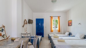 A room with a dining table, chairs, a TV, a blue door, and three beds with towels.