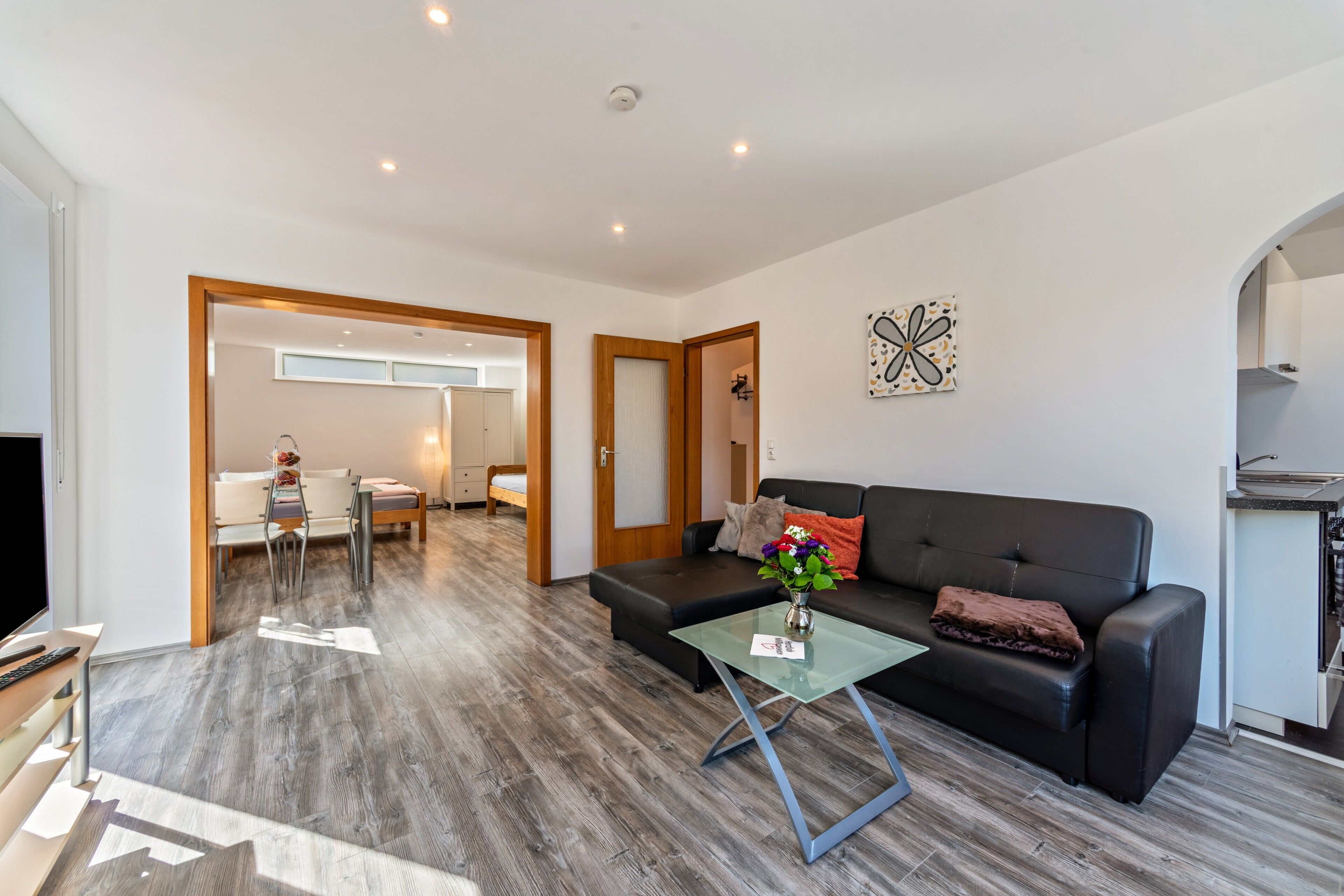 A modern living room with a sofa, coffee table, and a TV. There is a dining area with a table and chairs, and a kitchen area with a countertop.