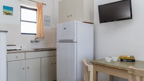 Ett kök med ett kylskåp, ett fönster med gardin, en diskho, ett träbord med böcker och en platt-TV som är monterad på väggen.