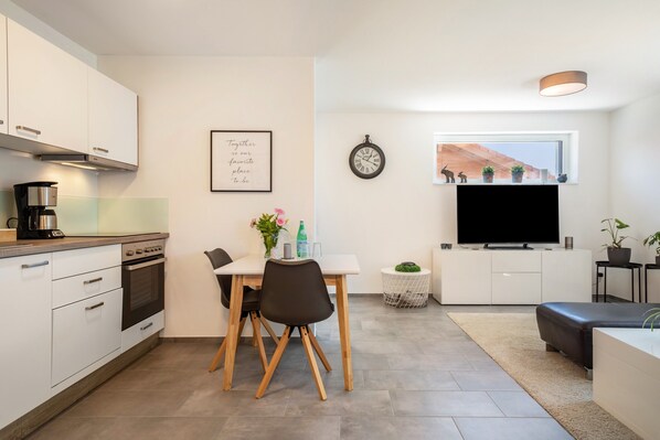 Une cuisine moderne avec des meubles blancs, une table à manger avec des chaises, une horloge, un téléviseur et une fenêtre donnant sur un beau paysage.
