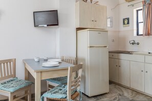 Une petite cuisine équipée d’un réfrigérateur, d’une table à manger avec des chaises, d’un téléviseur fixé au mur et d’une fenêtre avec des rideaux.