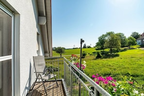 A balcony with a chair, a small table, and a view of a green landscape with trees and flowers.