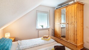 Una camera da letto con soffitto spiovente, un armadio in legno, un letto con cuscino blu e una finestra con persiane.