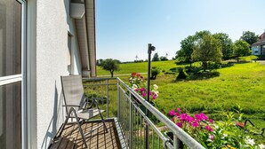 Un balcone con una sedia, una ringhiera e vista su un prato verde con alberi e fiori.