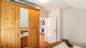 Una camera da letto con un armadio in legno, un letto con un asciugamano arrotolato e una valigia, e una cucina visibile sullo sfondo.