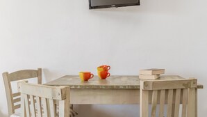 A wooden table with two orange cups and a book, a television mounted on the wall, and two wooden chairs. - Holiday Home '3 Kypri Seaview Perasma Studio' with Sea View, Wi-Fi and Air Conditioning (Kypri)