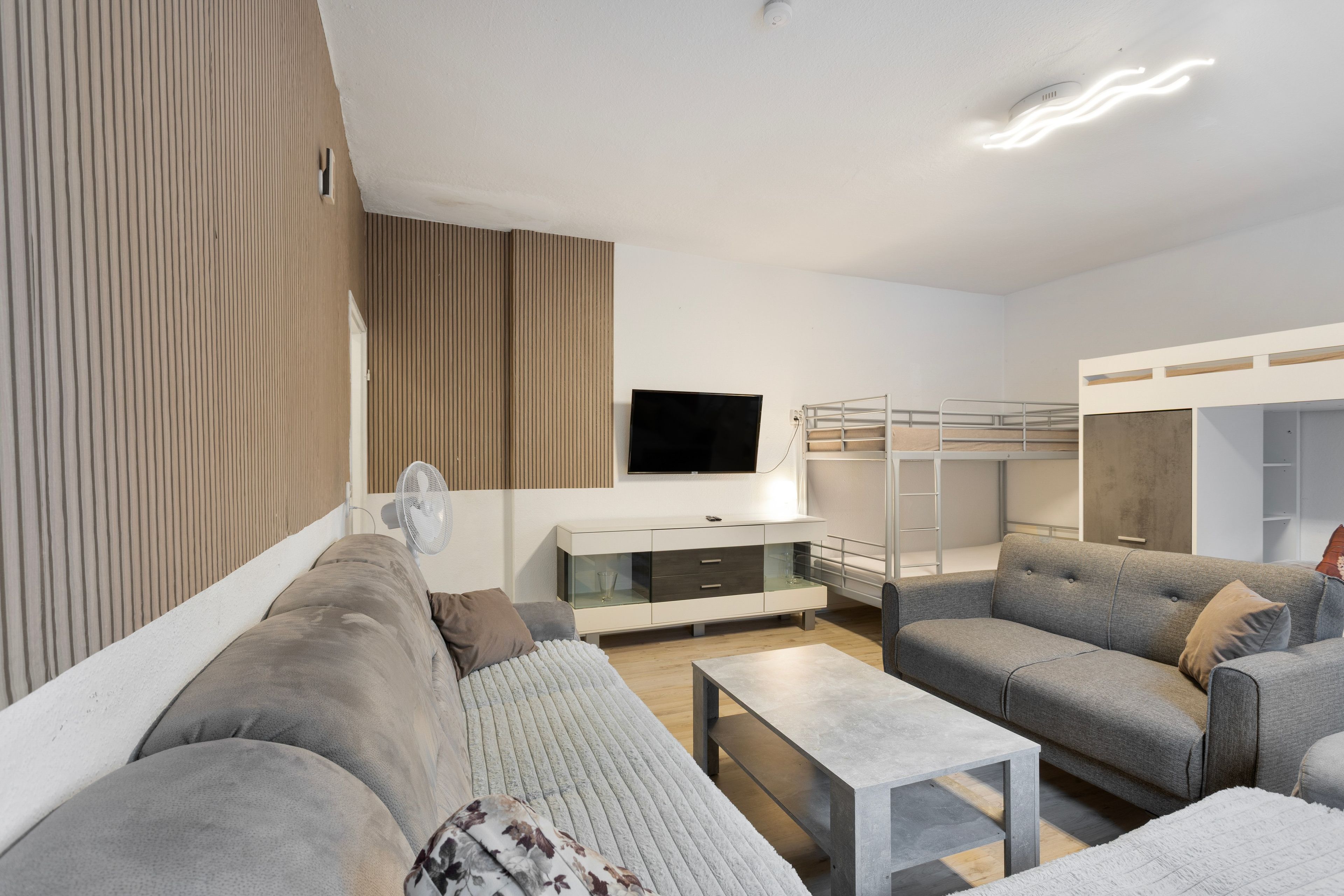 Un salon moderne avec un canapé, un fauteuil et une petite table. Un téléviseur est fixé au mur, et on aperçoit un lit superposé au fond.