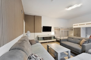 Een moderne woonkamer met een bank, fauteuil en een kleine tafel. Een televisie is aan de muur bevestigd en er staat een stapelbed op de achtergrond.