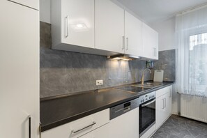 Une cuisine moderne avec des meubles blancs, un plan de travail noir, un four encastré et une fenêtre ornée de rideaux légers.