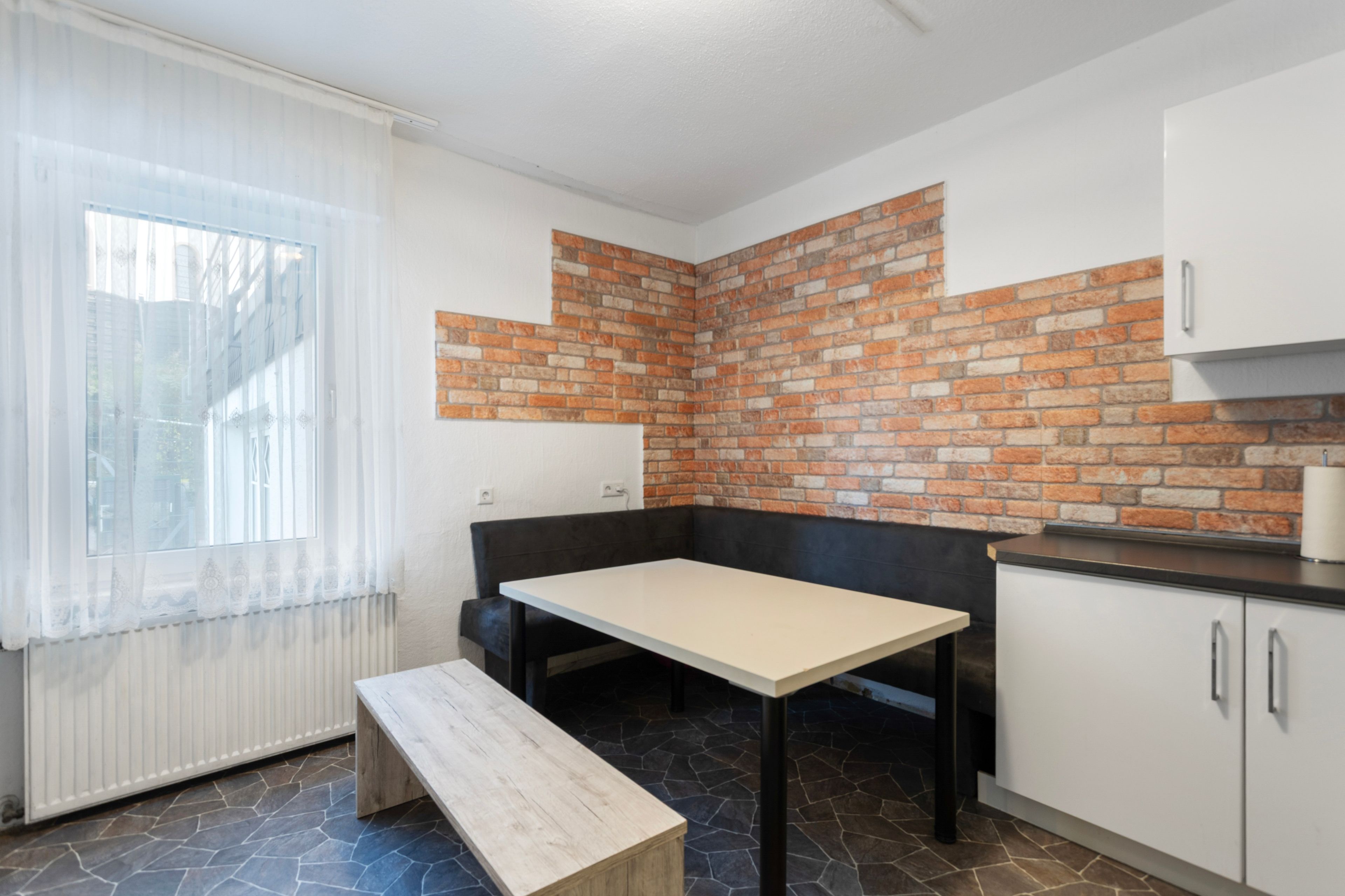 Una cocina con pared de ladrillo decorativa, una mesa de comedor, un banco y una ventana con cortinas translúcidas.