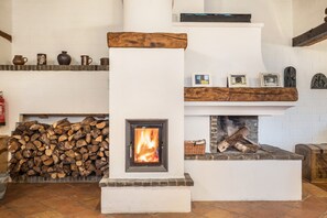Une cheminée avec un feu visible, une pile de bois de chauffage et une étagère ornée d’objets décoratifs.