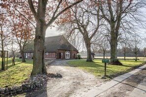 Une maison de campagne avec un toit de chaume, entourée d’arbres et accessible par un chemin de gravier.
