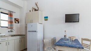 A compact kitchen with a refrigerator, a dining table with chairs, a television mounted on the wall, and a window with a curtain.