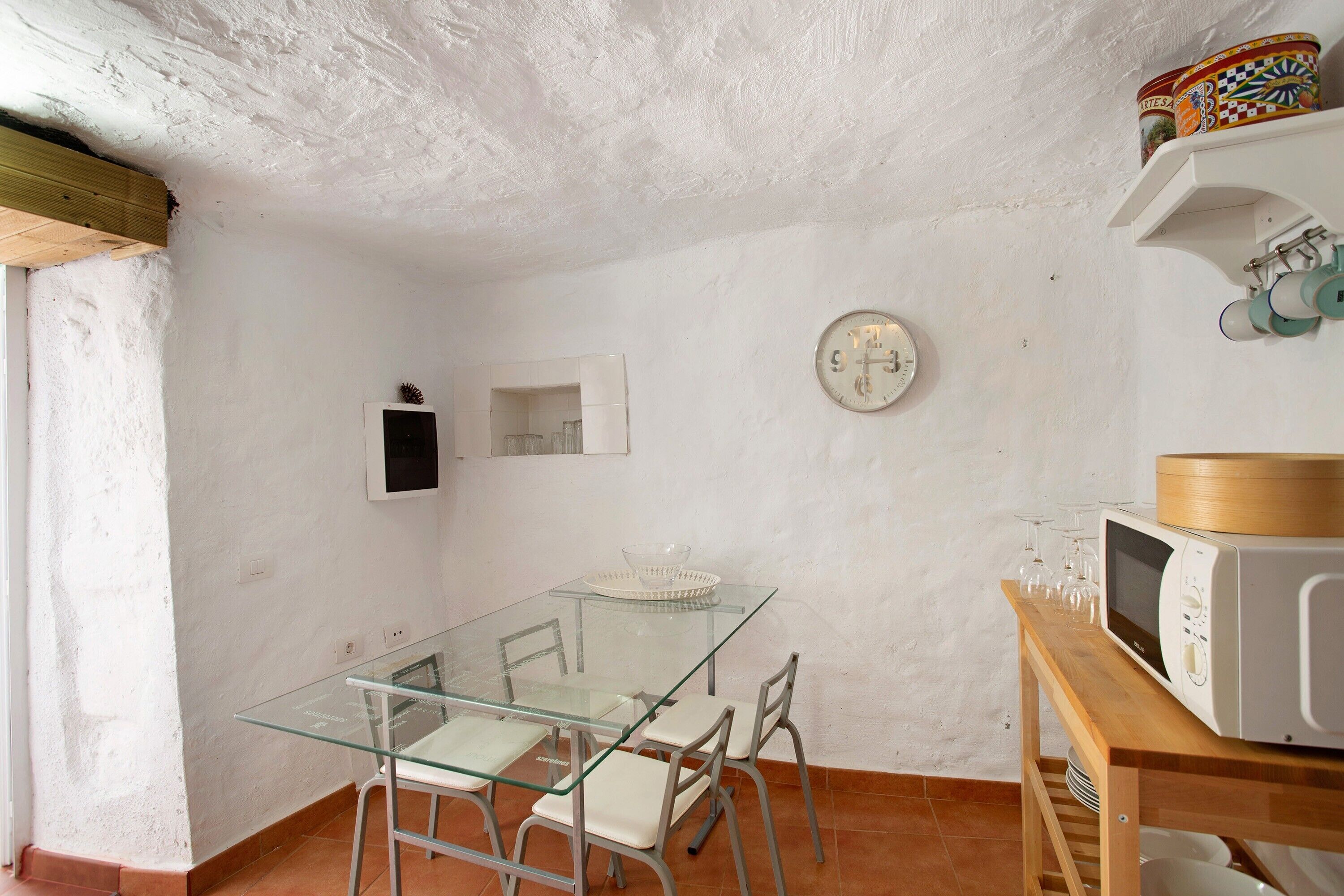 Cocina compacta con mesa de vidrio, sillas blancas, microondas y un reloj en la pared.