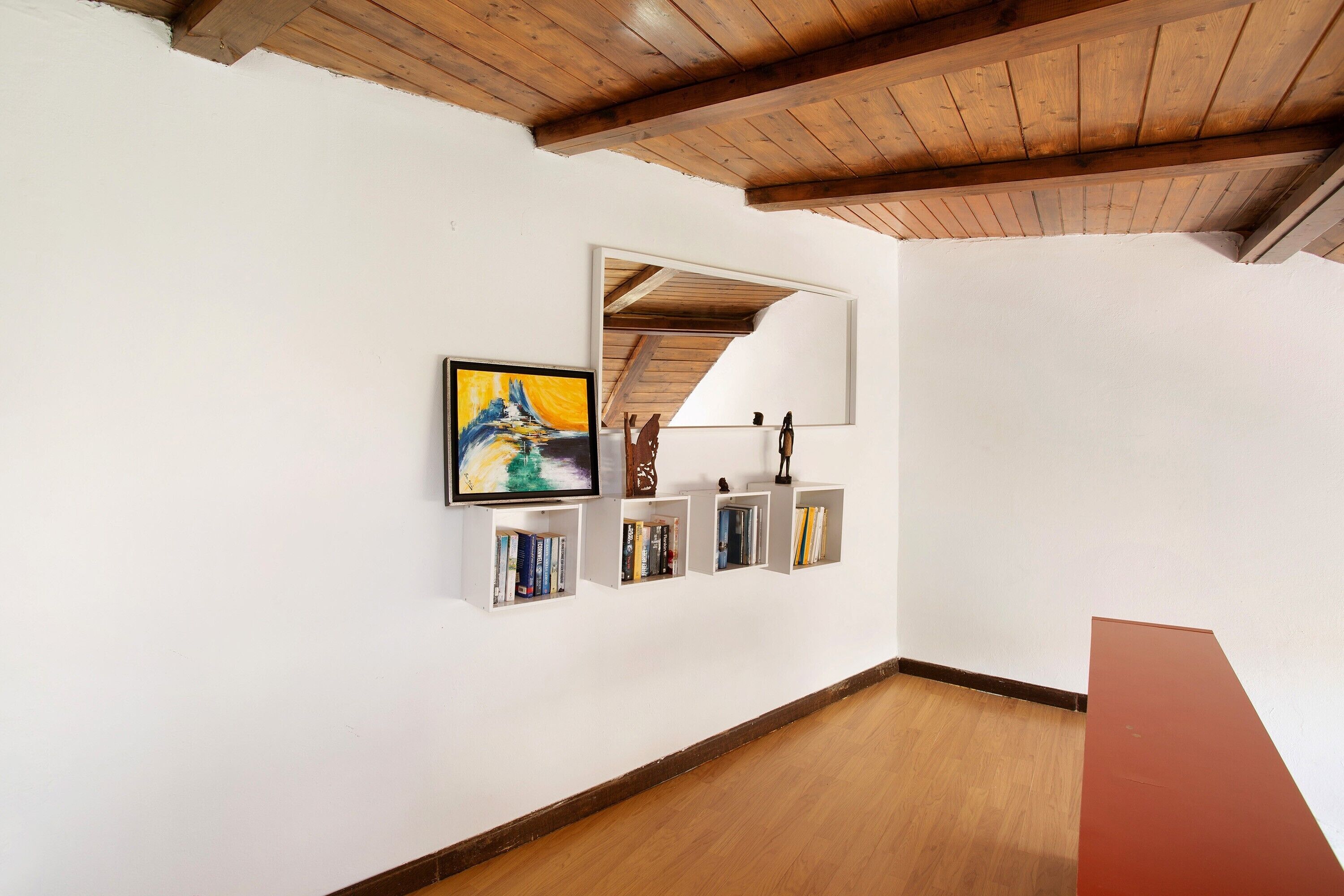 Una habitación con techo de madera, paredes blancas y suelo de madera. Hay un cuadro en la pared y un espejo encima.