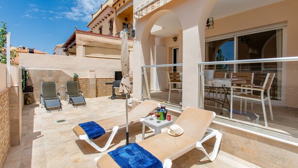 Un espace extérieur ensoleillé avec deux chaises longues, une petite table avec une boisson et une porte vitrée donnant sur un balcon.