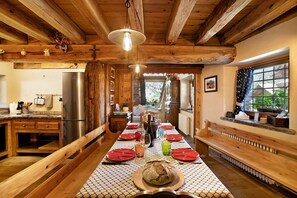 A wooden interior with a long dining table set for four, a refrigerator, and a window with curtains.