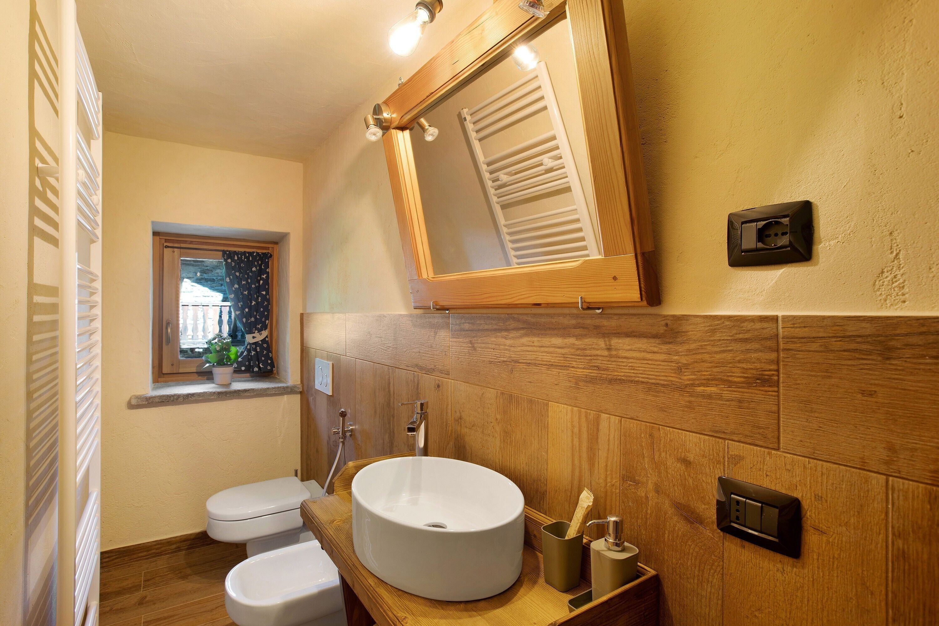A bathroom with a wooden vanity, a round sink, a toilet, a window with a view, and a mirror.