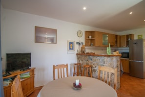 Un coin repas avec une table ronde, des chaises en bois, un téléviseur posé sur un meuble et un espace cuisine avec des placards en bois et un réfrigérateur.