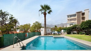 A swimming pool with a palm tree, a small white building, and a multi-story building in the background. - Apartment 'Prealsa Benalmádena' with Sea View, Shared Pool and Private Terrace (Benalmádena)