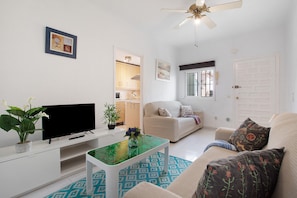 A living room with a flat-screen TV, a coffee table with a vase, a sofa with patterned cushions, a ceiling fan, and a door with a window.