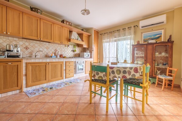 Une cuisine avec des meubles de rangement en bois, une table à manger avec des chaises, un sol carrelé et un appareil de climatisation.