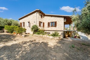 Una casa de piedra con balcón, rodeada de vegetación y un camino de entrada de grava.