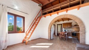 A spacious room with a stone floor, a wooden staircase, a kitchen area, and a large window with curtains.