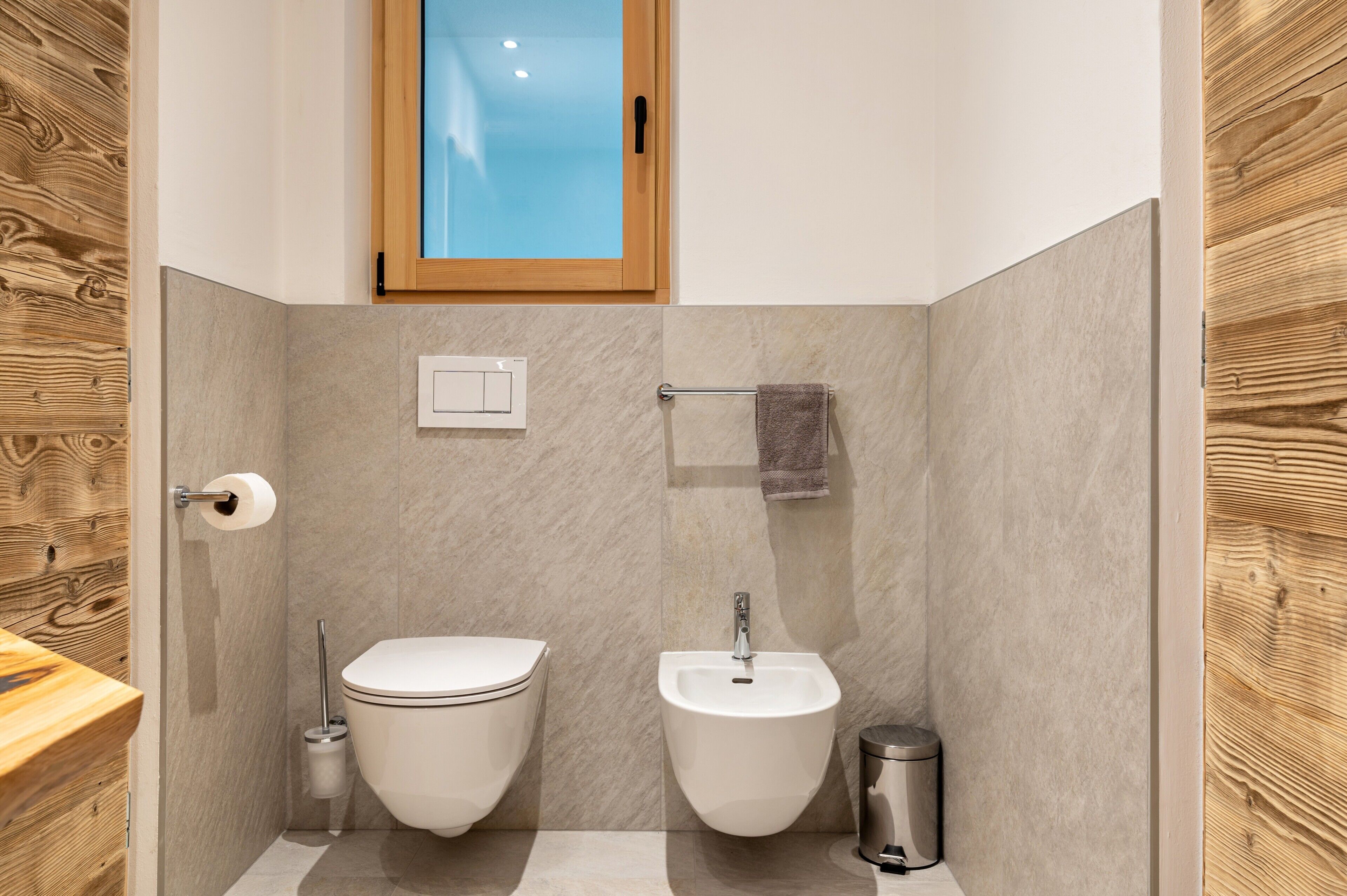 Un baño moderno con inodoro, bidé y lavamanos, paneles de madera en la pared y una ventana.