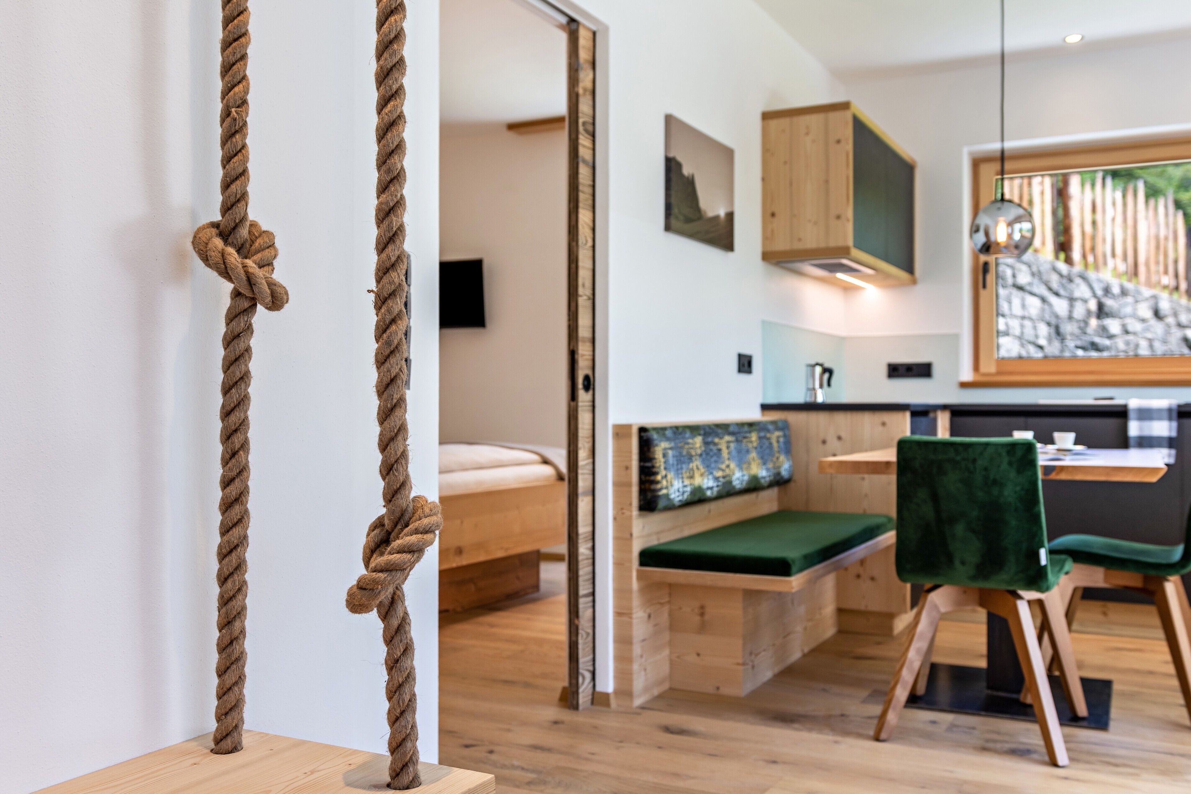 Un interior moderno con piso de madera, una cama con cojín verde, una zona de comedor con mesa y sillas, y una cocina con gabinetes de madera.