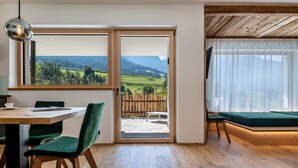 A room with a wooden floor, a dining table with chairs, a sofa, a large window with a view of a green landscape, and a sliding glass door. - Holiday Apartment "Chalet Kasch Glück" with Mountain View, Private Terrace & Wi-Fi (Fiè allo Sciliar)