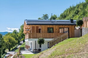 Une maison moderne en bois, dotée d’un balcon, d’une petite dépendance attenante et offrant une vue sur une colline.