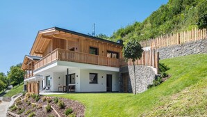 Una casa moderna de dos pisos con balcón de madera, un muro de contención de piedra y un jardín cubierto de césped.
