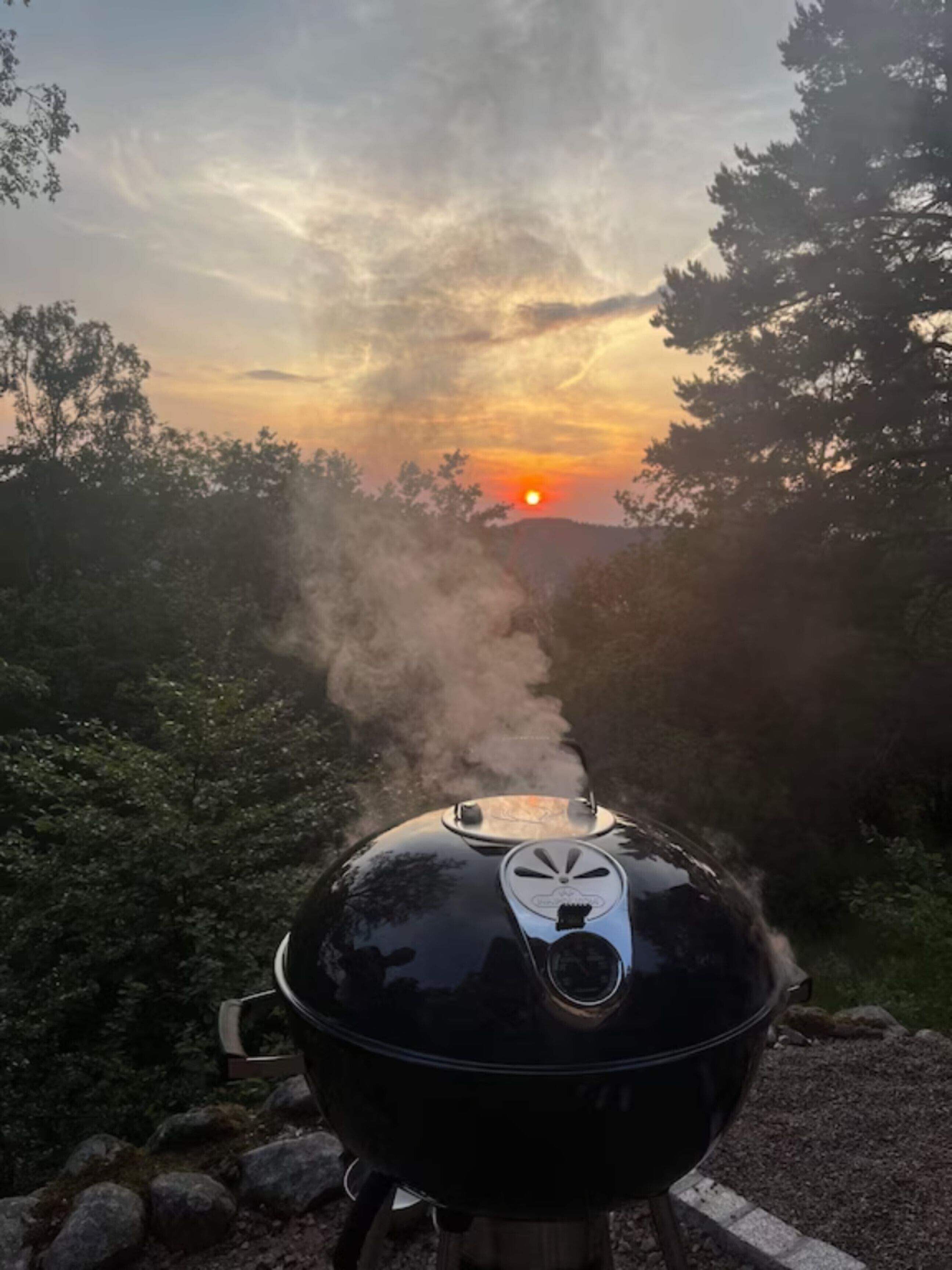 En sort grill med ventilationsrist, placeret på en stengulv, omgivet af træer og en solnedgang i baggrunden.