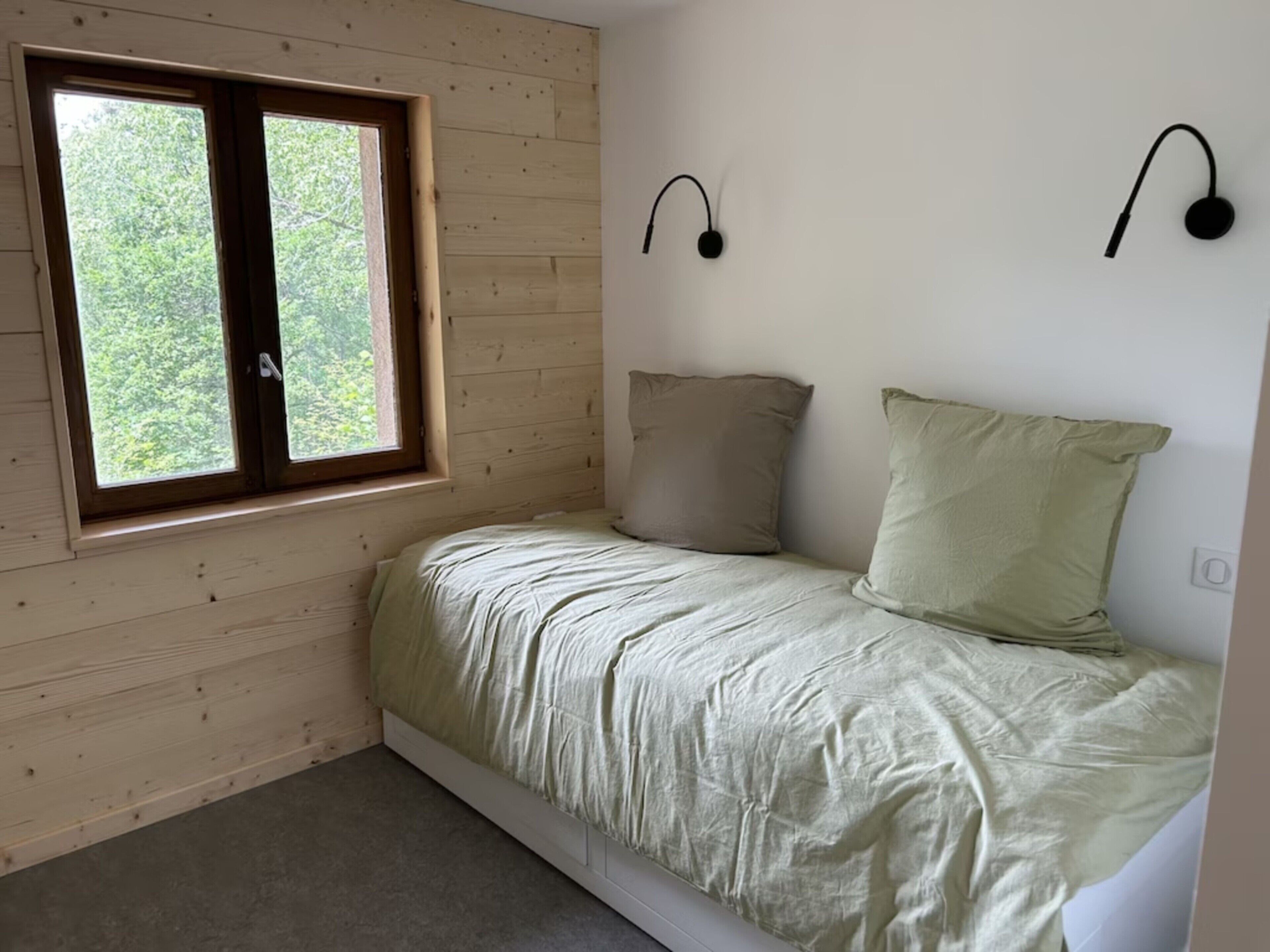 A single bed with a headboard, a window with a view of trees, and two wall-mounted light fixtures.