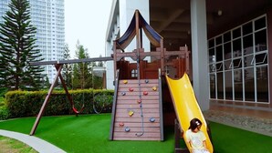 Une aire de jeux avec un toboggan et un mur d’escalade, entourée d’herbe verte et d’arbres.