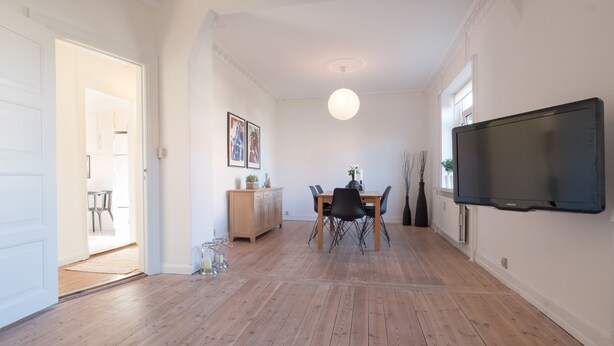 A spacious living room with a wooden floor, a dining area with a table and chairs, a flat-screen TV mounted on the wall, and a door leading to another room. - Spacious 2-Bed Apartment in Aalborg (Aalborg)