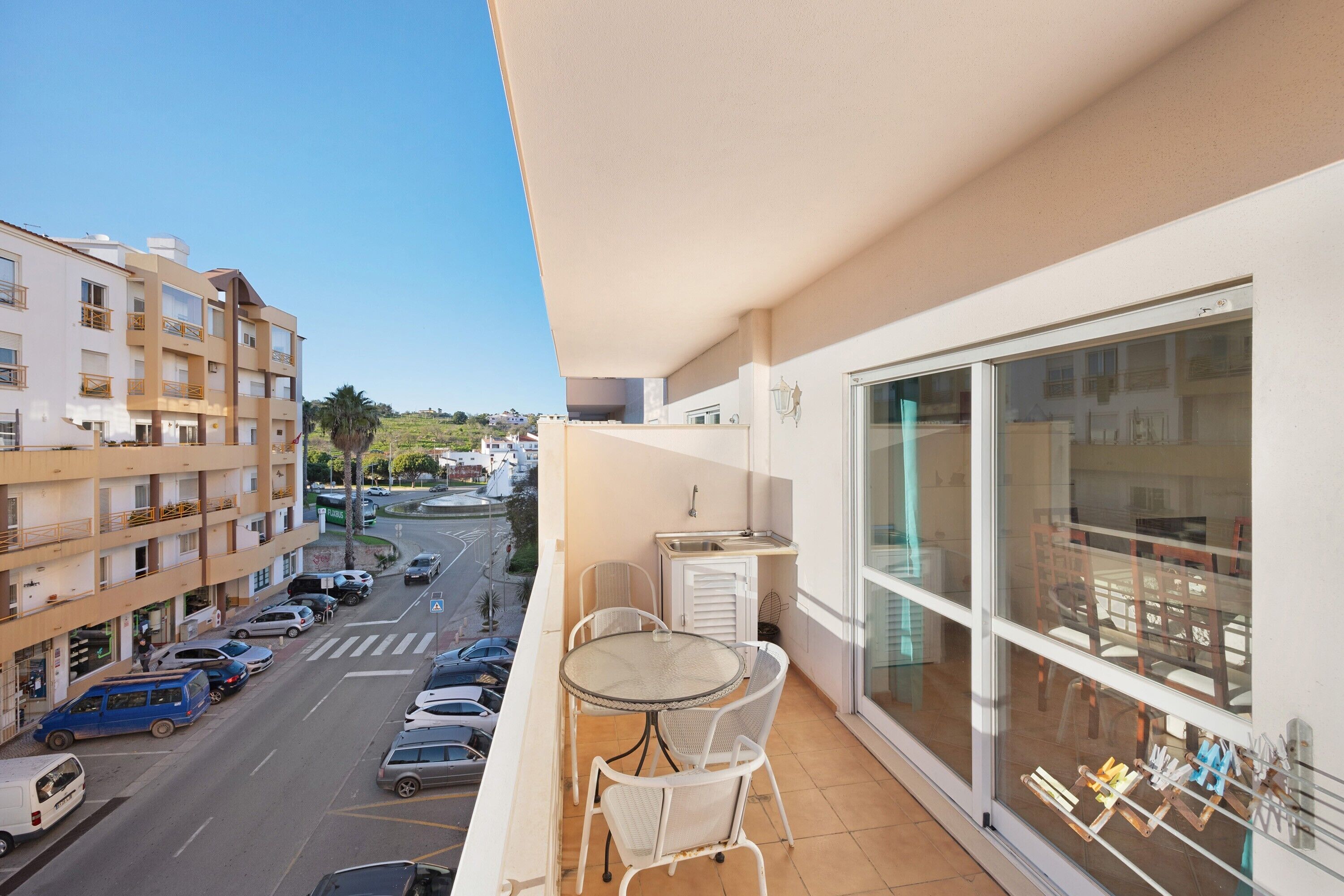 A balcony with a table and chairs, a view of a street with parked cars, and a building with balconies.